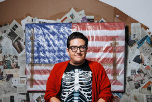 Artist Just Favela poses in front of an American Flag in a skeleton tee and red sweater