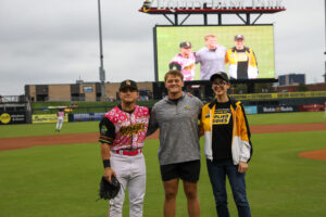 Ian Stevens, a Wichita State physical education student, stands on the field at Riverfront Stadium with a Wichita Wind Surge player and College of Applied Studies Dean, Dr. Jennifer Friend. The stadium scoreboard behind them shows the group’s photo during Credit Union of America’s Educators Night.