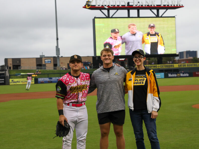 Ian Stevens, a Wichita State physical education student, stands on the field at Riverfront Stadium with a Wichita Wind Surge player and College of Applied Studies Dean, Dr. Jennifer Friend. The stadium scoreboard behind them shows the group’s photo during Credit Union of America’s Educators Night.
