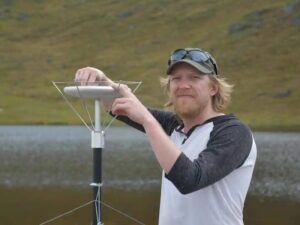 Photograph of Dr. Clay Prater standing in a field, standing next to radar used to track animals. Behind Dr. Prater is a body of water that is nearly indistinguishable from the grass surrounding it. Dr. Prater is smiling at the camera and wearing a ball cap with sunglasses resting on the brim.