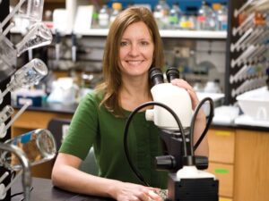 Photograph of Dr. Erika Geisbrecht sitting behind a microscope in a laboratory setting. Dr. Geisbrecht is smiling at the camera with one hand resting on the microscope's ocular lens.