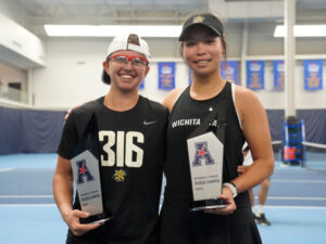 Xin Tong Wang and Giorgia Roselli pose at blue tennis courts holding their championship trophies that say Women's Tennis Doubles Champion 2025