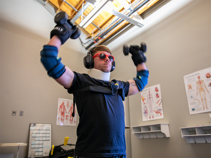 A physical therapy student at Wichita State University tests a homemade geriatric-simulation suit with wrist weights, dumbbells, elbow braces, sunglasses, earmuffs and a neck brace