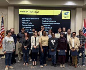 Group of student awardees, some of them holding their certificates and certificate holders. They are joined, on the right, by Graduate Dean Coleen Pugh, and on the left, by Associate Dean Enrique Navarro. On the back, there is a screen listing the names of some of the nominees under the title "Congratulations". On the right hand, the US flag. On the left hand, the Wichita flag.
