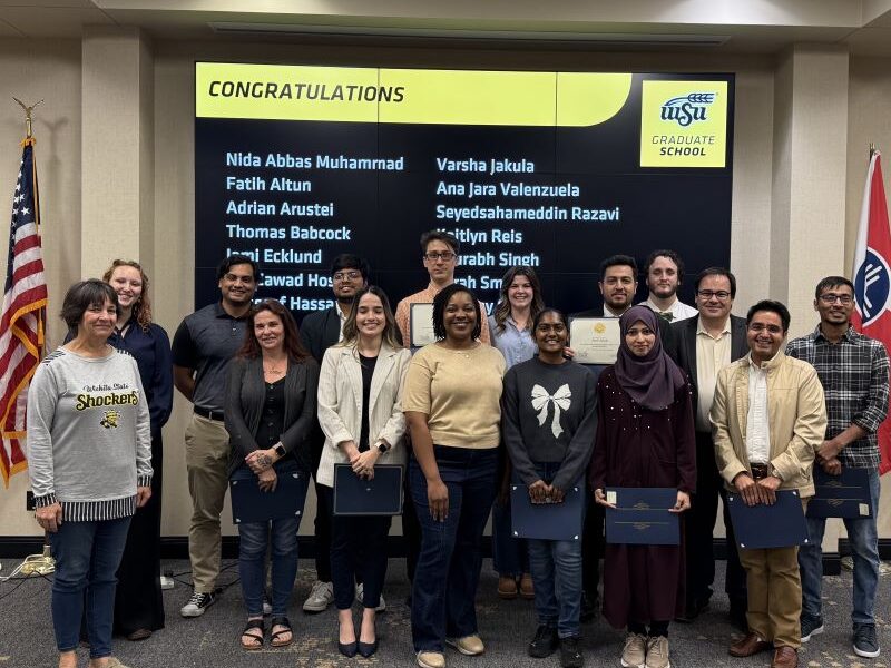 Group of student awardees, some of them holding their certificates and certificate holders. They are joined, on the right, by Graduate Dean Coleen Pugh, and on the left, by Associate Dean Enrique Navarro. On the back, there is a screen listing the names of some of the nominees under the title "Congratulations". On the right hand, the US flag. On the left hand, the Wichita flag.