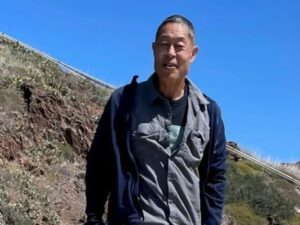 A man with close cut grayish hair standing near a mountainous region wearing a gray button down shirt and navy zipper sweatshirt.
