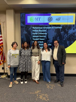 Five people are standing indoors in front of a large digital display screen and smiling for a group photo. To the left side of the image, there is an American flag. The people are dressed in semi-casual to business-casual clothing. The floor is carpeted, and the setting appears to be a presentation or conference room.