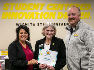 Three smiling people standing in front of a Student Centered Innovation Driven sign. Woman in middle holding a certificate.