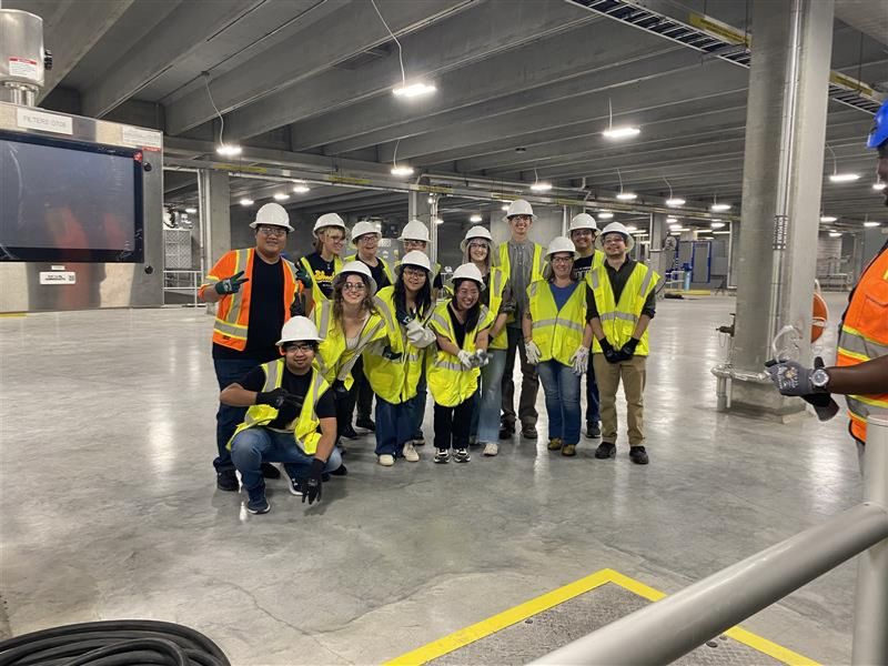 A group of students wearing hard hats and safety vests pose in a water treatment plant with pipes and machinery.