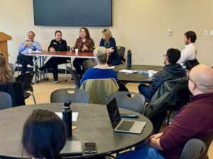 Four faculty members sit at a table at the front of a room during a panel for December's New Faculty On-Ramp program. Other faculty sit around tables listening.