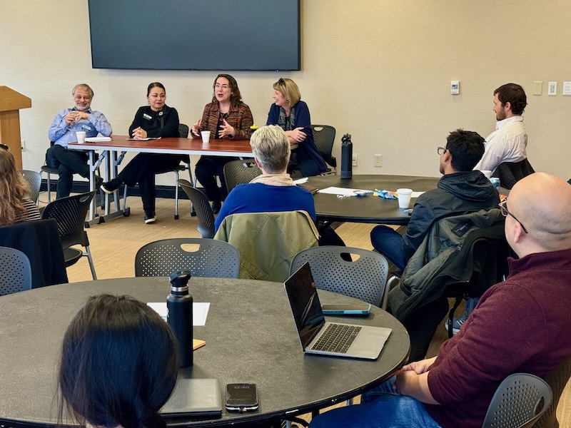 Four faculty members sit at a table at the front of a room during a panel for December's New Faculty On-Ramp program. Other faculty sit around tables listening.