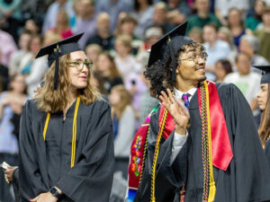 A male student in graduation regalia waves to the crowd at commencement as a female student in graduation regalia talks to him