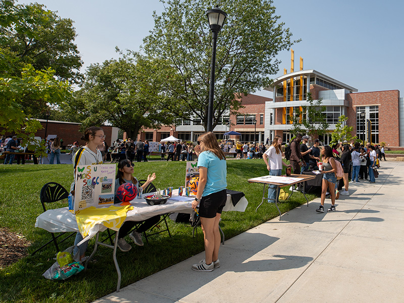 A student visits a women's soccer table in front of the Rhatigan Student Center with many students in the background visiting other student organization tables