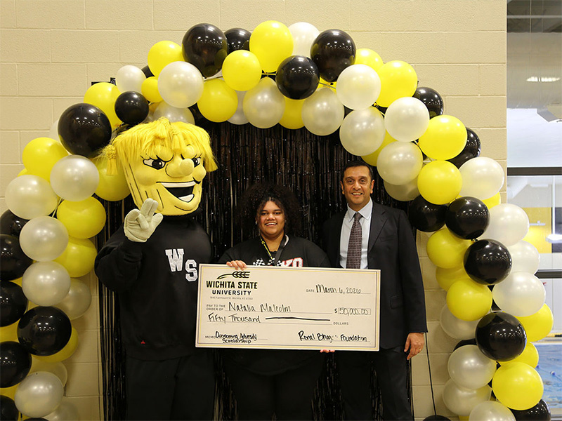 Natalia Malcolm poses with WuShock and Ronal Bhagat while holding onto the $50,000 scholarship check in front of white, black and yellow balloons and a black curtain