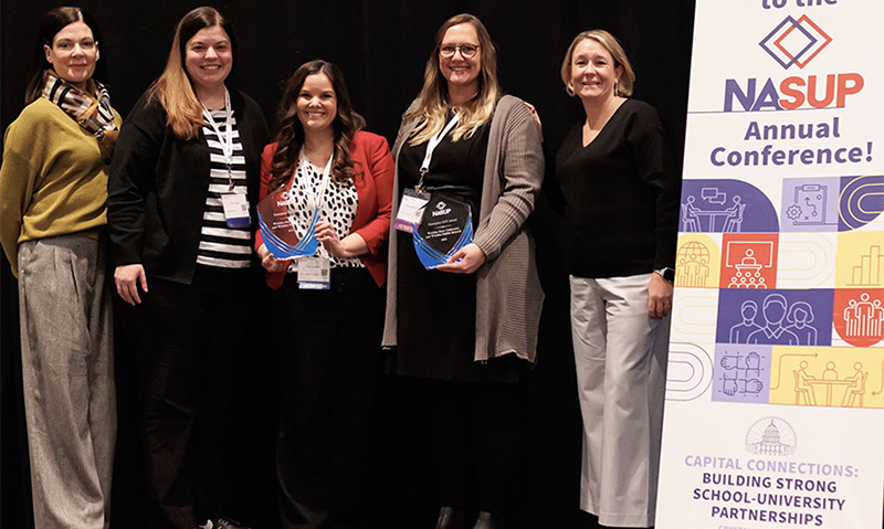 Representatives from Wichita State University and Wichita Public Schools hold up awards next to a sign for the NASUP annual conference and the 2026 Exemplary School-University Partnership Award from the National Association for School-University Partnerships (NASUP)