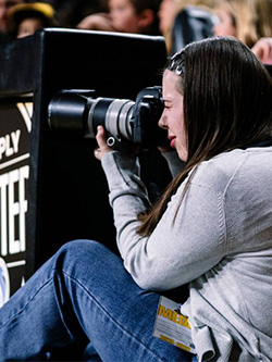 Ellery Prothro sits on the sidelines during a men's basketball game, shooting photography with a camera in her hands and up to her face
