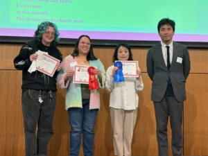 Four people stand on a stage. The first three, Jack, person wearing black with pale green hair, Necia, a person with long hair in a multicolored pastel sweater, and Yumi Foster, who is wearing a white shirt with tan pants, hold up certificates with white, red, and blue ribbons. The fourth person is a man in a grey suit with a thin black tie wearing a name tag.
