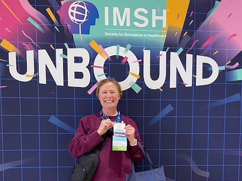 Jennifer Thornberry poses with her badge in front of wall art that says "IMSH: Society for Simulations in Healthcare. Unbound"