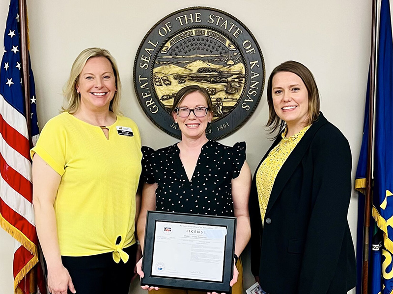 Three women stand in front of the Great Seal of the State of Kansas and U.S. and Kansas flags at the Kansas State Board of Education meeting. The woman in the center holds her framed teaching license while two Wichita State representatives stand beside her.