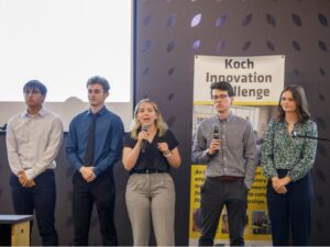 A team of five freshman students, three men and two women, deliver their elevator pitch to an audience at the 2025 Koch Innovation Challenge. They are standing in front of a banner with the words "Koch Innovation Challenge."