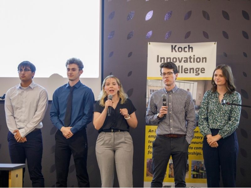 A team of five freshman students, three men and two women, deliver their elevator pitch to an audience at the 2025 Koch Innovation Challenge. They are standing in front of a banner with the words "Koch Innovation Challenge."