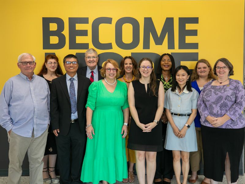 Hugo Wall School Faculty and Staff stand in front of a yellow wall with the word "BECOME" in large black letters.