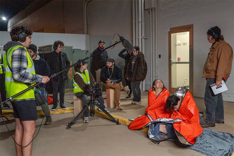 Students in thermal cloths sit on the ground acting as students with camera and audio equipment record the scene