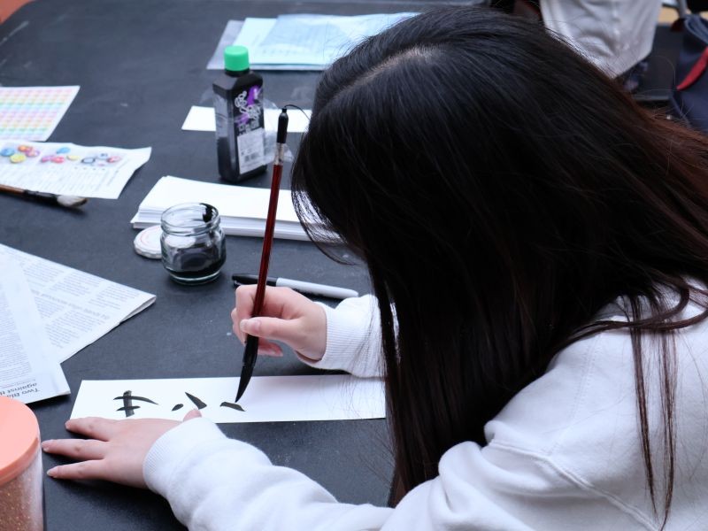 A girl with long dark hair is bent over a long, skinny sheet of paper and holding a traditional Japanese calligraphy brush in one hand, writing Japanese characters on the sheet.
