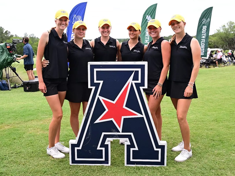 Members of the Shocker women's golf team pose behind the American Conference logo in a grassy field