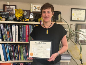 Moriah Beck holds up her AAAS Fellow plaque in front of a bookshelf full of various textbooks and research material