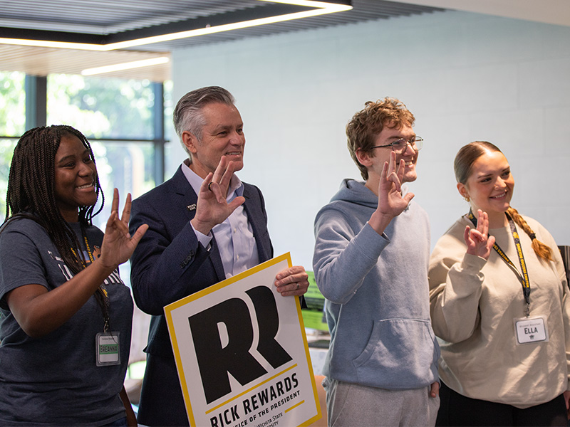 President Rick Muma poses with students while holding a Rick Rewards sign as they all hold up the Shockers hand sign