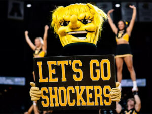 WuShock holds up a sign that says "Let's Go Shockers" with members of the Shocker cheer team in the background in Charles Koch Arena.