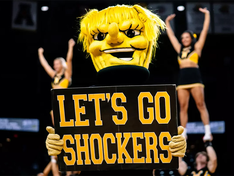 WuShock holds up a sign that says "Let's Go Shockers" with members of the Shocker cheer team in the background in Charles Koch Arena.