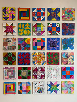 A grid of square quilts of different designs on a white wall.