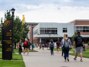 Students walking on campus, a sign with directions to various building sits off the path, and a building with the words Rhatigan Student Center on it is in the background.