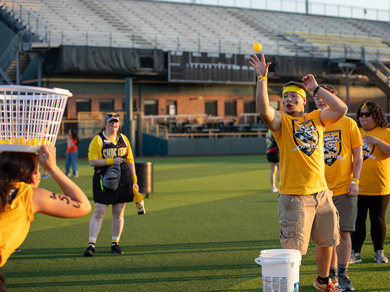 A student tosses a ball into a basket held by another student on top of their head.