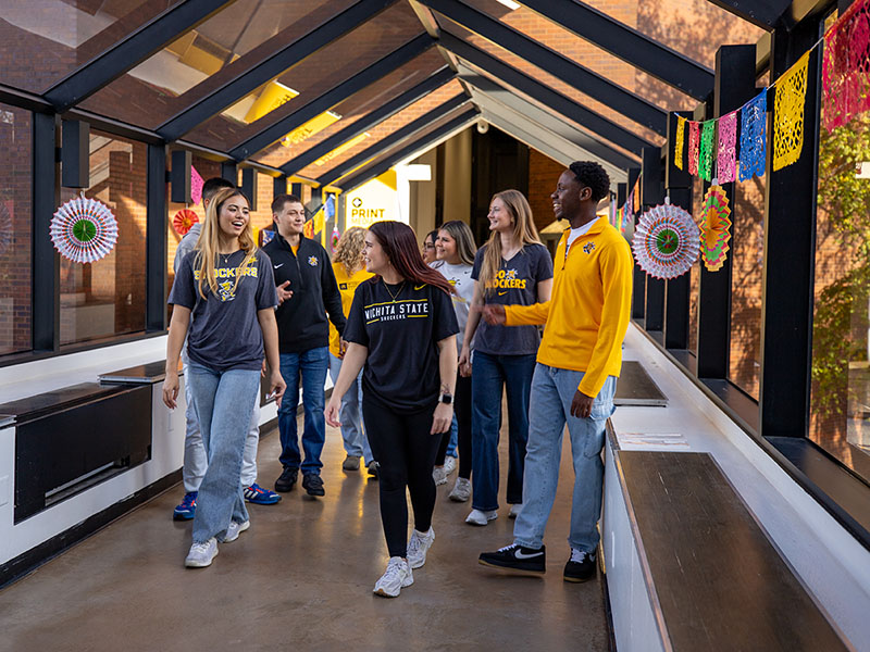 Students mingle in an overground walkway with various art pieces hanging from the wall. The walkway is open on the sides and ceiling with glass windows and support beams between them