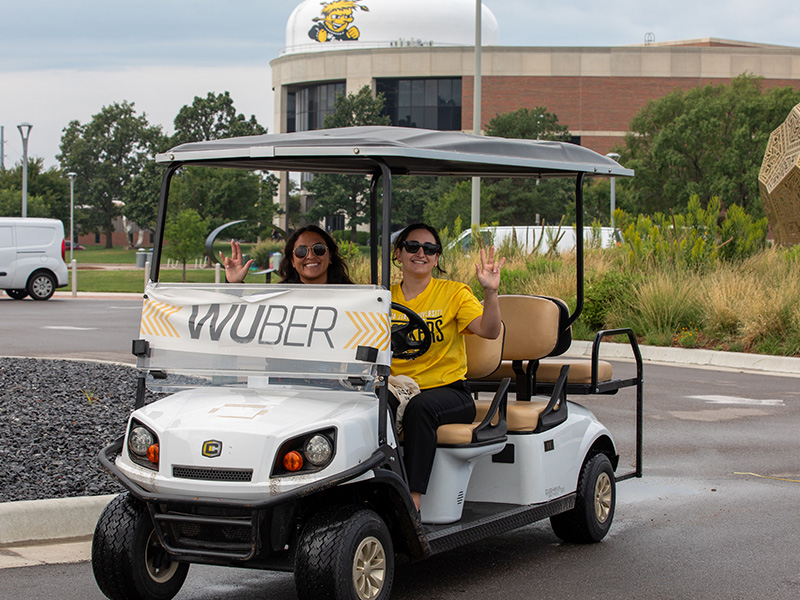 Shocker ambassadors wave to the camera as they drive a golf cart with a "Wuber" banner on the front.