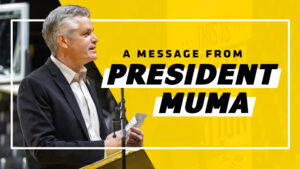 The text "A message from President Muma" on a yellow background that cuts the graphic in half. On the other half is Rick Muma speaking to a crown from a podium.