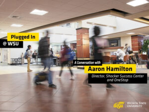 Students walk through a busy indoor campus commons area at Wichita State University. Several people are shown moving across the foreground with motion blur, while others sit and study at tables in the background. Overlay text reads "Plugged In @ WSU" and "A Conversation with Aaron Hamilton, Director, Shocker Success Center and OneStop." The Wichita State University logo appears in the lower-right corner. The image promotes a podcast episode featuring WSU Success Coaches and highlights student activity and engagement on campus.