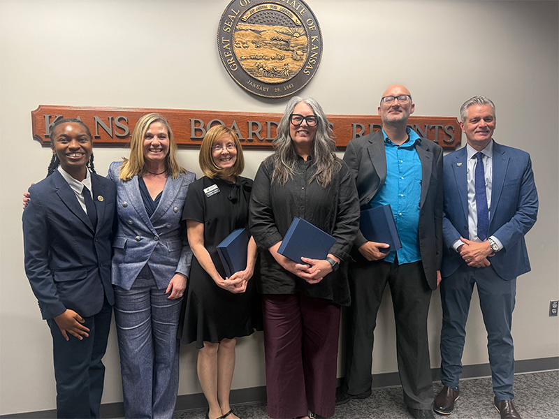 Winners of the Faculty/Staff of the Year award pose in front of a sign that says "Kansas Board of Regents" and the KBOR seal with the WSU president, provost and SGA president. Each staff/faculty member are holding a box containing their awards.