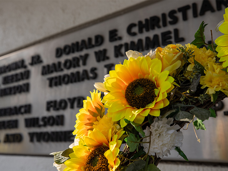 Sunflowers sit in front of the Memorial '70 sculpture. Behind the sunflowers are the list of names of the victims of the plane crash.