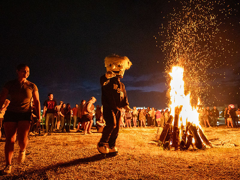 WuShock points to the camera standing next to a bonfire. Students are in the background.
