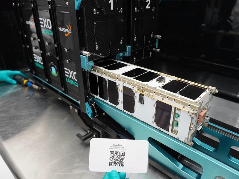 The SNAPPY CubeSat before its launch sits on a table with a student holding up a card with a QR on it next to it
