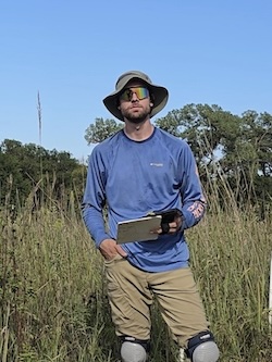 Gavin Page standing in tall prairie, looking at the camera while holding a clipboard.