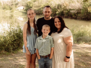Kristen Knott with her husband James, daughter Kenzie and son Brantley in a grassy field with a pond in the background.