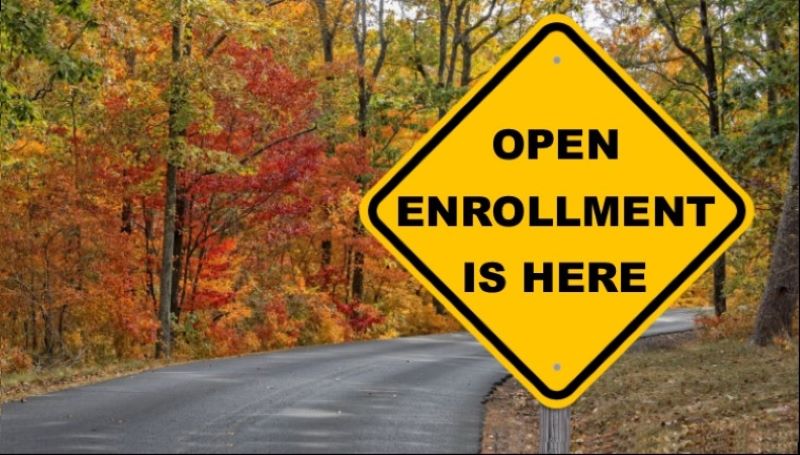 A road sign with the words Open Enrollment Ahead is pictured on a country road that is surrounded on both sides by trees with fall foliage.