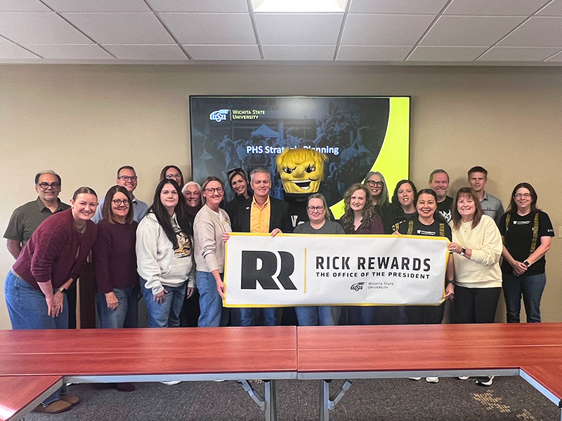 President Rick Muma and WuShock pose with members of the Department of Public Health Sciences holding up a banner that says "Rick Rewards."